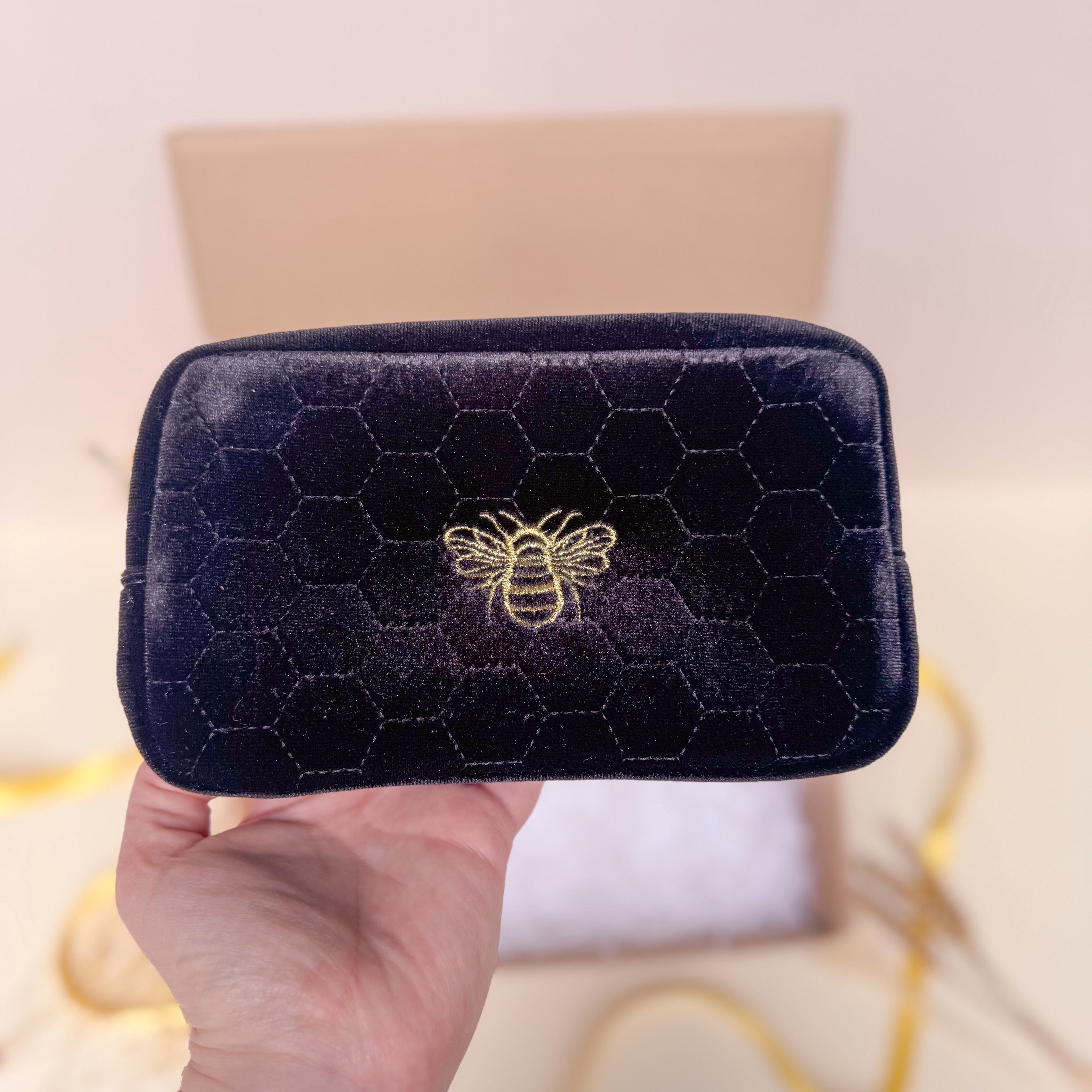 Black quilted velvet makeup bag with bee emblem held by a hand against a blurred background