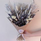 A hand holding a dried flower bouquet wrapped in brown kraft paper. The bouquet features a mix of white, silver, and grey dried grasses and flowers, with a round white sticker on the wrapper that reads 'WILLOW COTTAGE FLOWERS'.