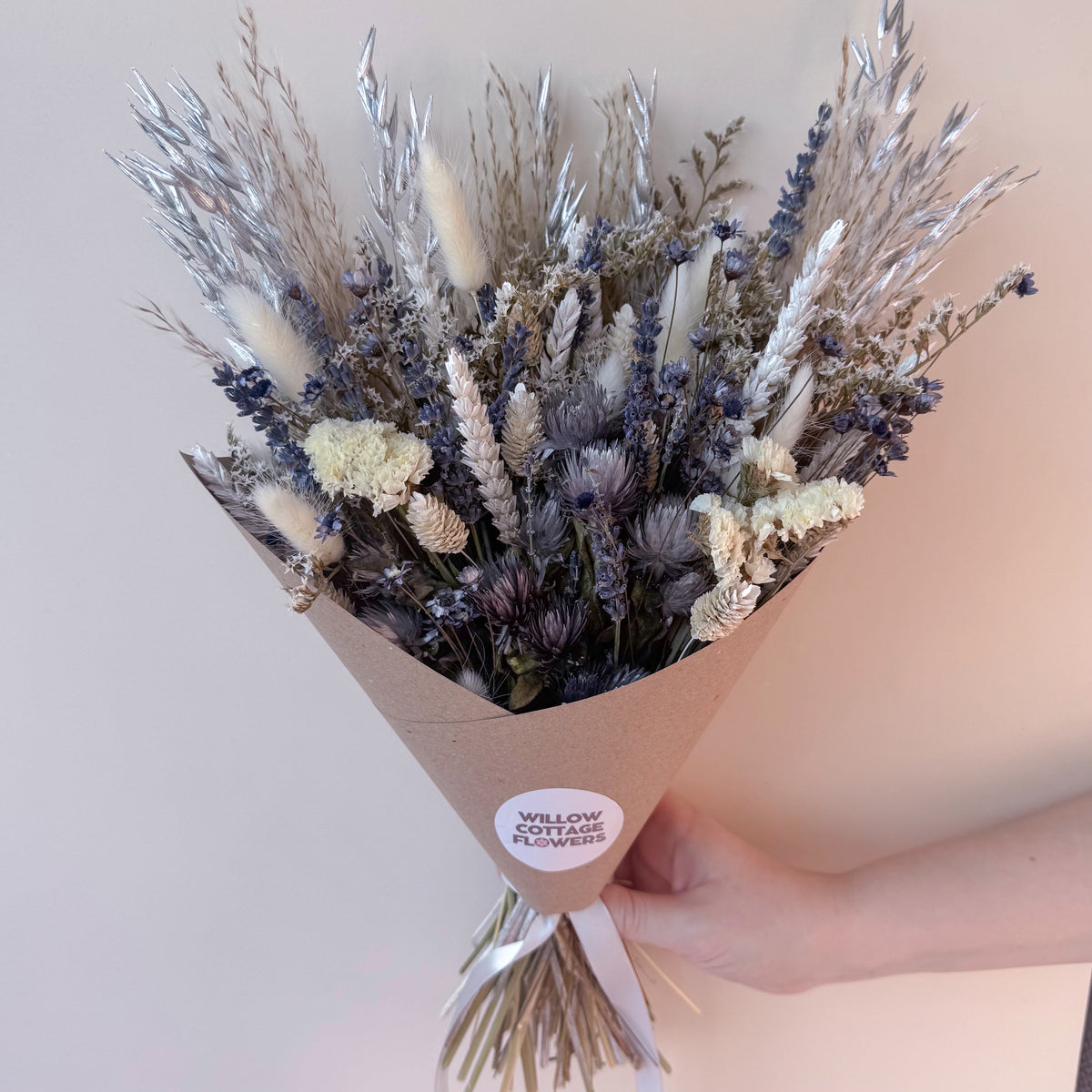 A hand holding a dried flower bouquet wrapped in brown kraft paper. The bouquet features a mix of white, silver, and grey dried grasses and flowers, with a round white sticker on the wrapper that reads 'WILLOW COTTAGE FLOWERS'.