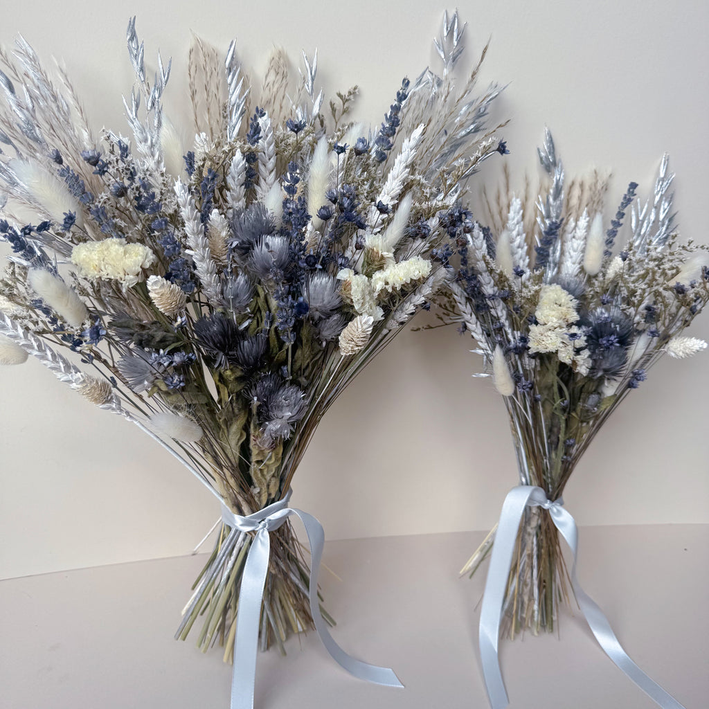 Two dried flower bouquets with silver, white, and grey botanicals tied with white satin ribbons, displayed against a plain beige background.