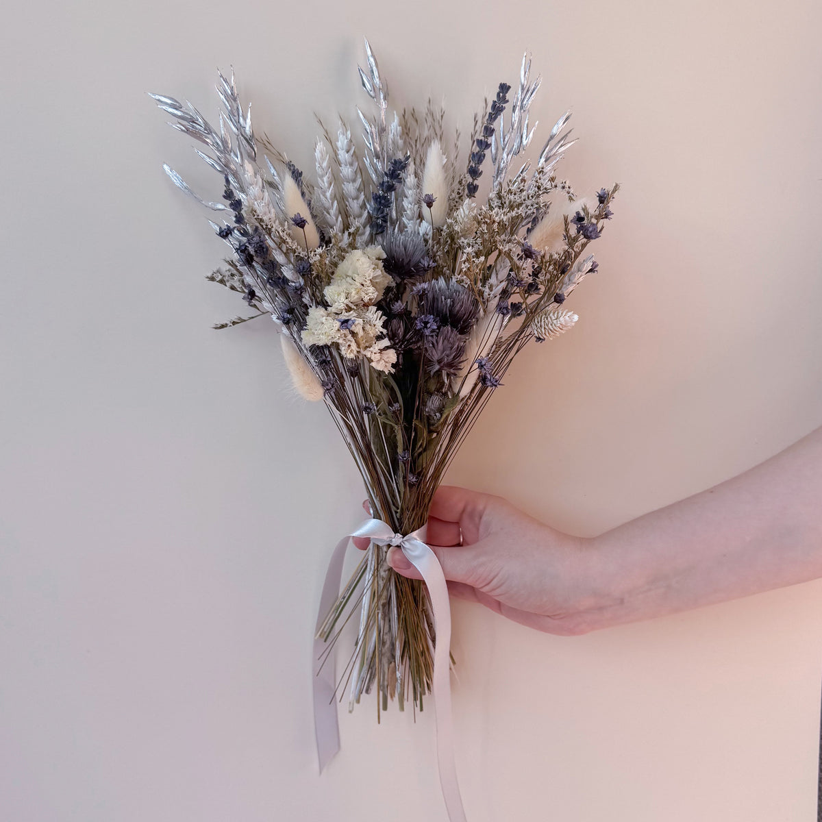 A hand holding a dried flower bouquet with lavender, wheat, and white filler stems tied with a pale grey ribbon, set against a solid beige background.
