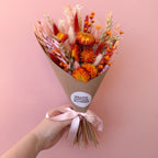 A hand holding a dried flower bouquet wrapped in brown kraft paper with a pink ribbon. The bouquet features vibrant orange, red, and beige dried flowers, set against a solid pink background.