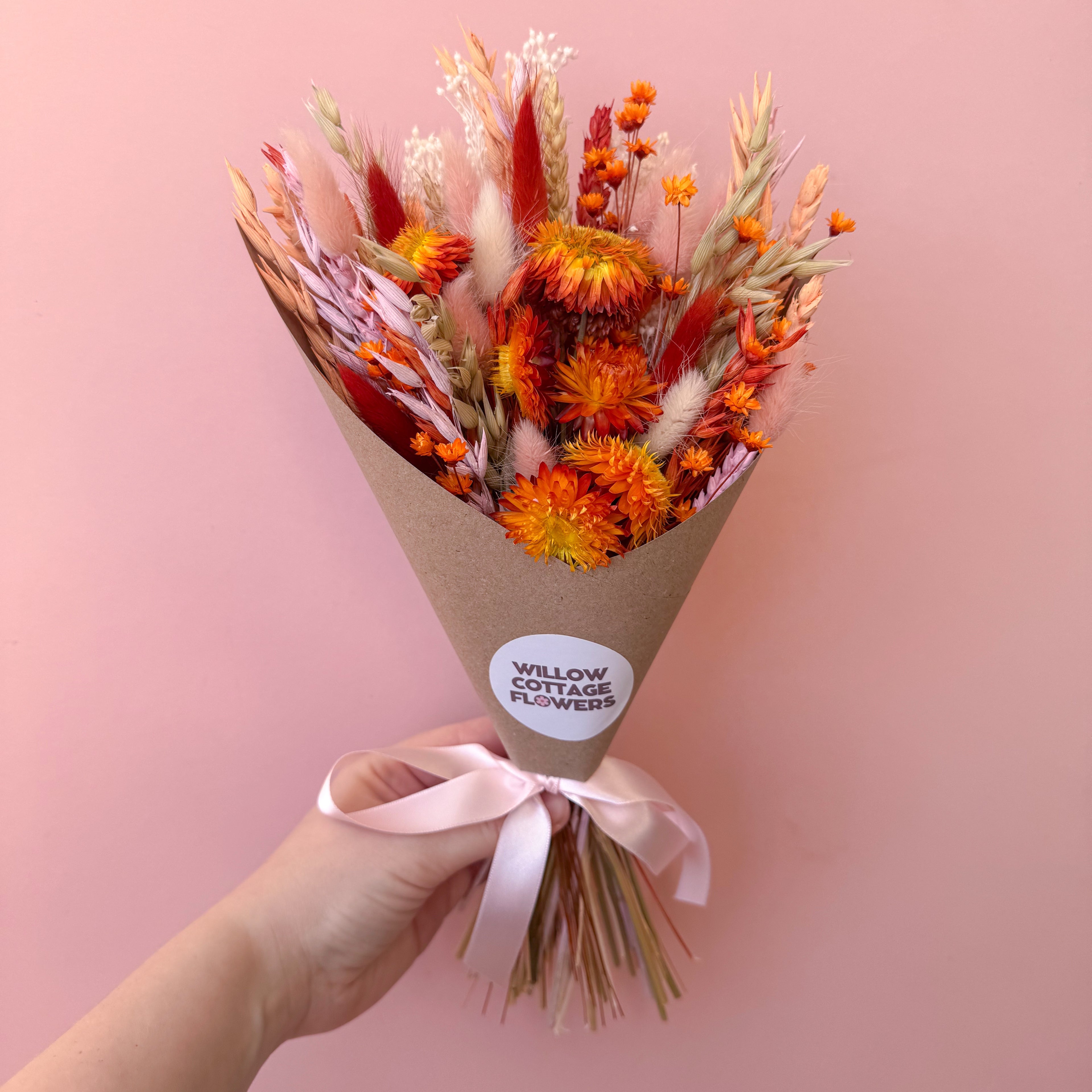 A hand holding a dried flower bouquet wrapped in brown kraft paper with a pink ribbon. The bouquet features vibrant orange, red, and beige dried flowers, set against a solid pink background.