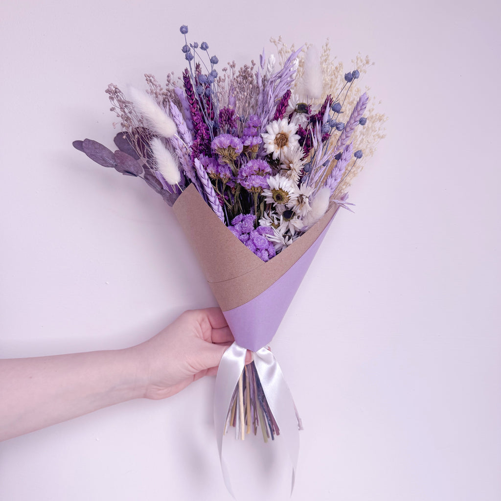 A hand holding a dried flower bouquet wrapped in brown kraft paper and light purple paper with a white ribbon, set against a solid pale pink background.