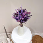 A bouquet of dried purple and violet flowers with bunny tails arranged in a small white speckled ceramic vase, placed on a round white stool against a plain beige wall.