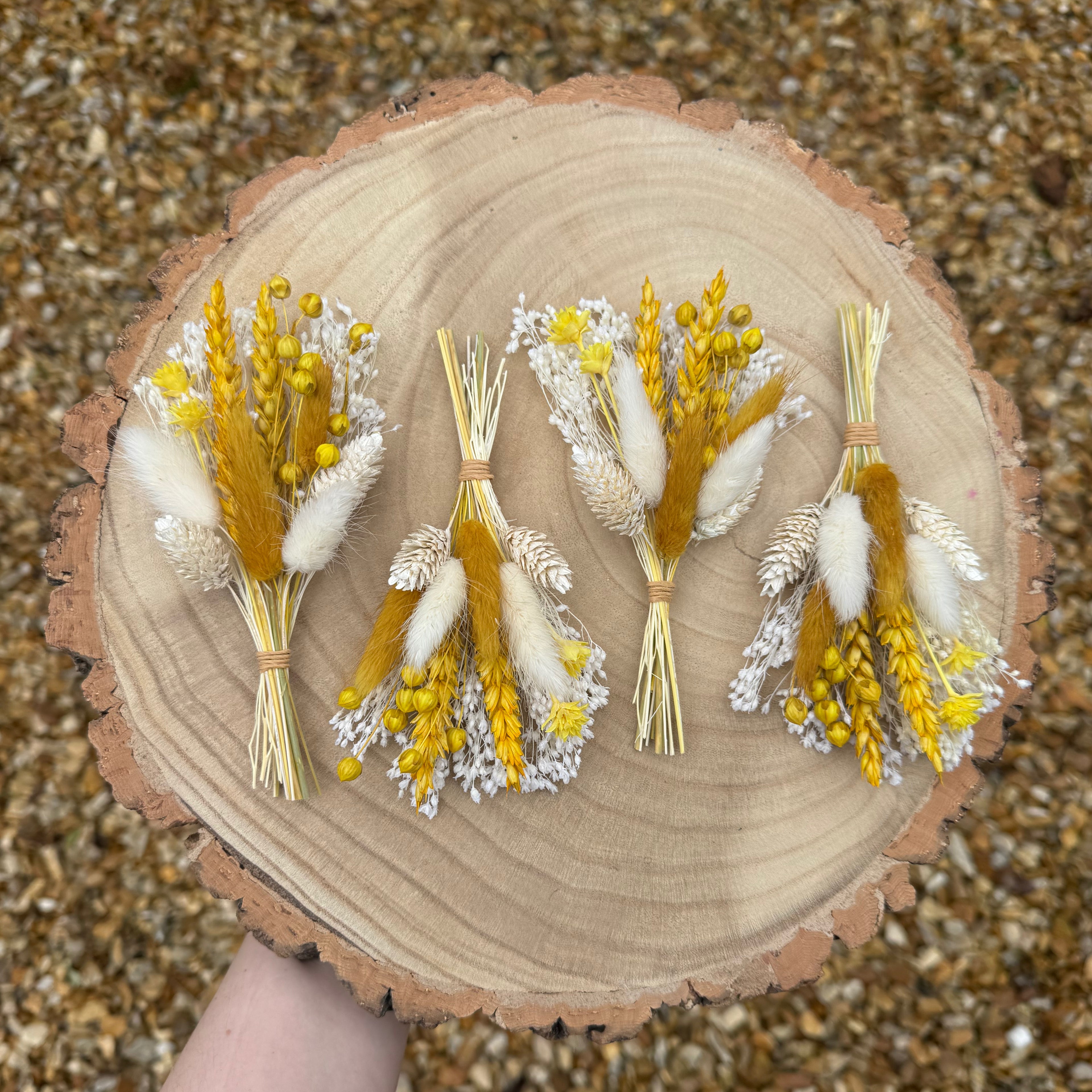 Four small dried flower buttonholes arranged on a round natural wood slice. A hand is visible holding the wood slice, and the background is gravel.