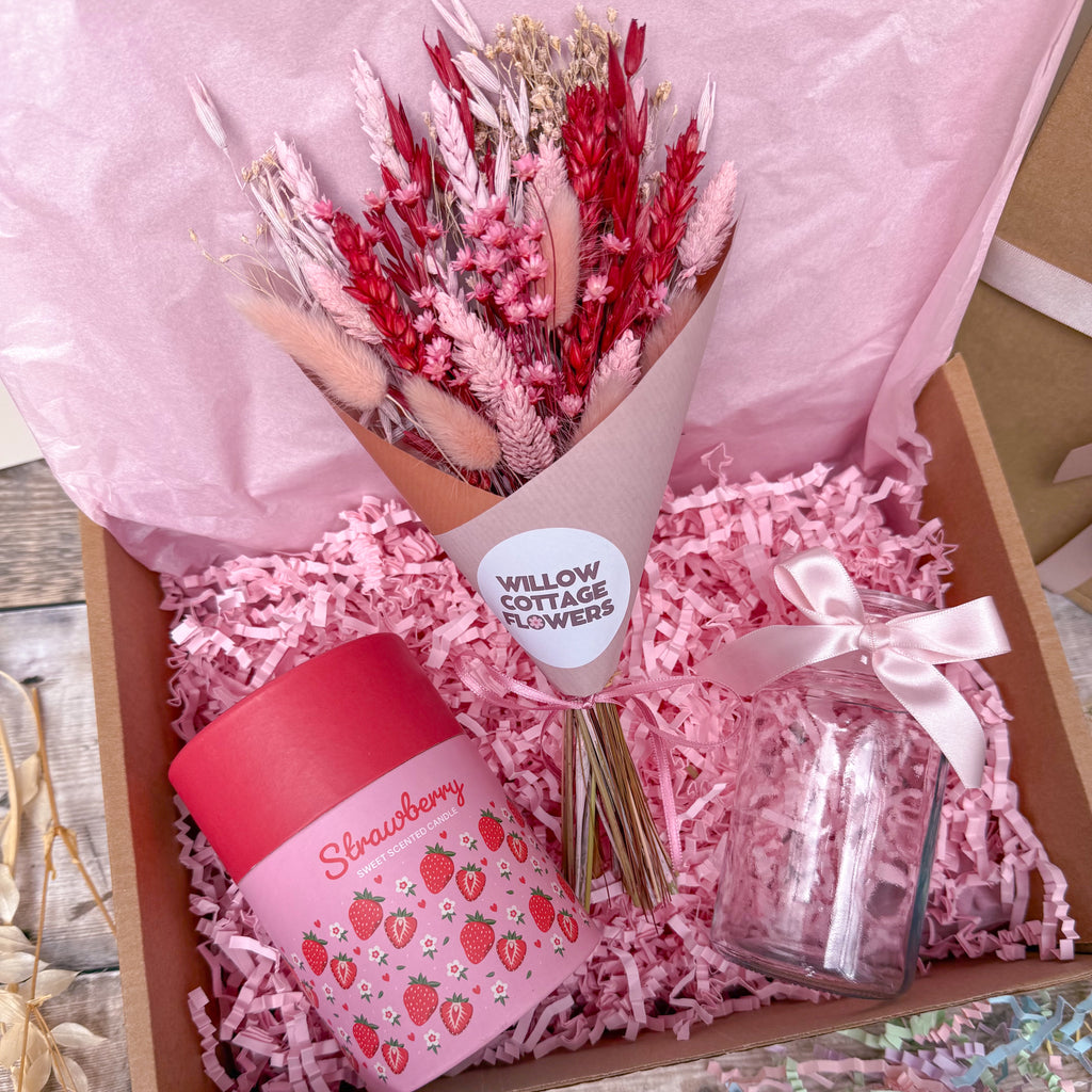 A pink gift box arrangement with a bouquet of dried red and pink flowers wrapped in tissue paper, a pink strawberry-themed candle in a red container, a clear glass jar tied with a pale pink ribbon, and pink shredded paper filler inside a brown box.