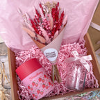 A pink gift box arrangement with a bouquet of dried red and pink flowers wrapped in tissue paper, a pink strawberry-themed candle in a red container, a clear glass jar tied with a pale pink ribbon, and pink shredded paper filler inside a brown box.
