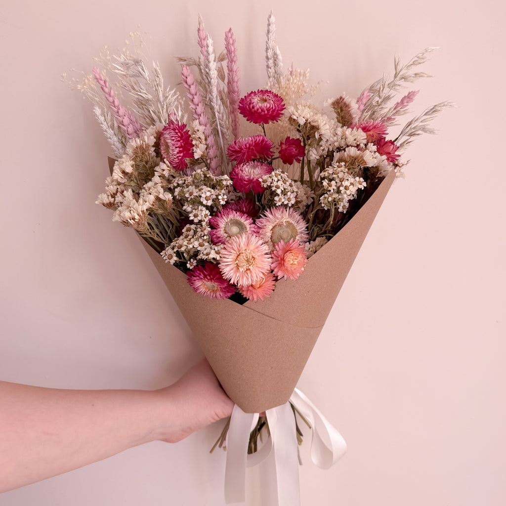Bouquet of daisy flowers wrapped in brown paper held by a hand against a light pink background