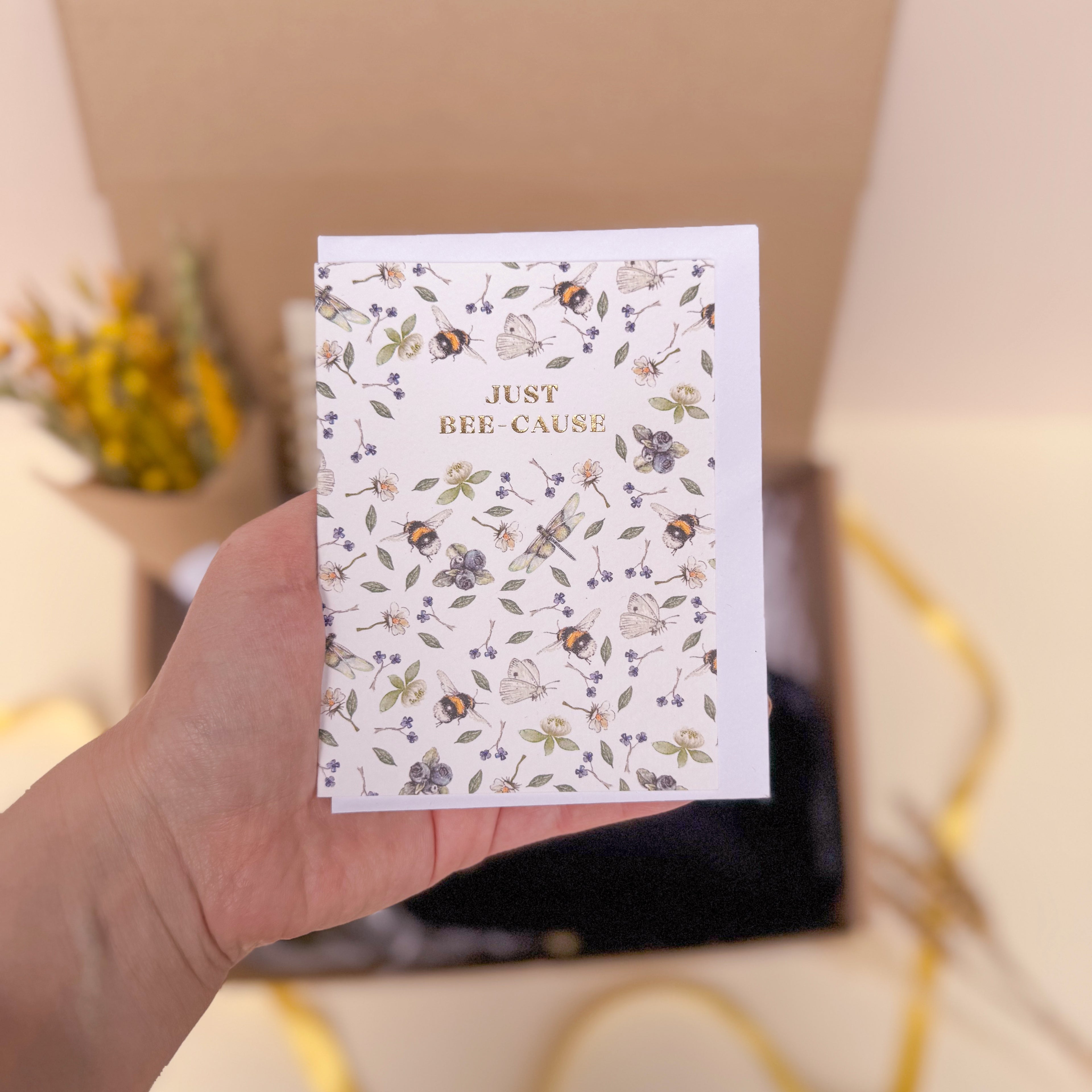 Hand holding a card with floral design and 'JUST BEE-CAUSE' text.