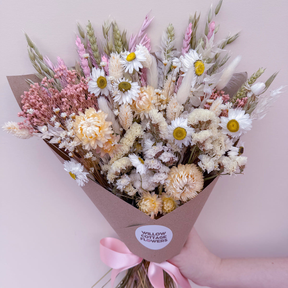 A dried flower bouquet wrapped in brown paper with a pink ribbon and a round label reading 'Willow Cottage Flowers,' held by a person's hand against a pale pink background.