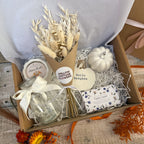 An autumn-themed gift box filled with cosy home decor items, including a dried flower bouquet in brown packaging, a white ceramic pumpkin, a candle, a glass vase with a white ribbon and a bar of luxury soap, all arranged in white shredded paper inside a cardboard box.