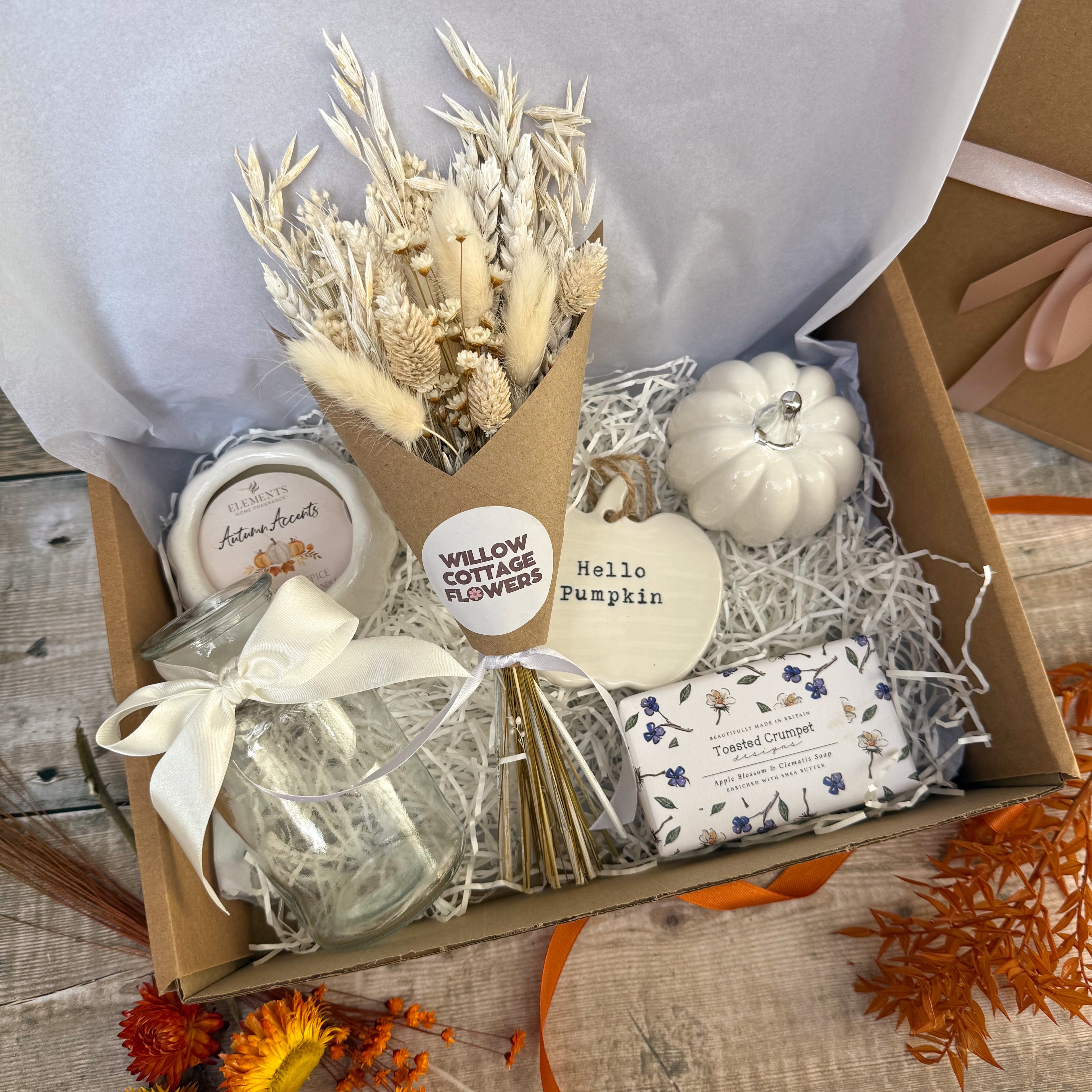 An autumn-themed gift box filled with cosy home decor items, including a dried flower bouquet in brown packaging, a white ceramic pumpkin, a candle, a glass vase with a white ribbon and a bar of luxury soap, all arranged in white shredded paper inside a cardboard box.