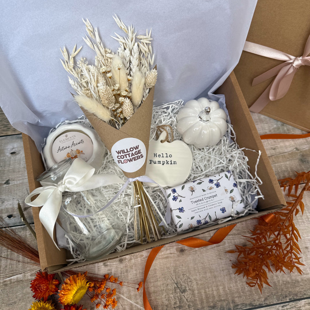 A curated gift box filled with autumn-themed items, including a dried flower bouquet in a kraft paper gift wrap, a small white decorative pumpkin, a wrapped bar of soap, a pumpkin shaped candle and a wrapped item with a 'Hello Pumpkin' tag, arranged in shredded paper inside an open cardboard box.