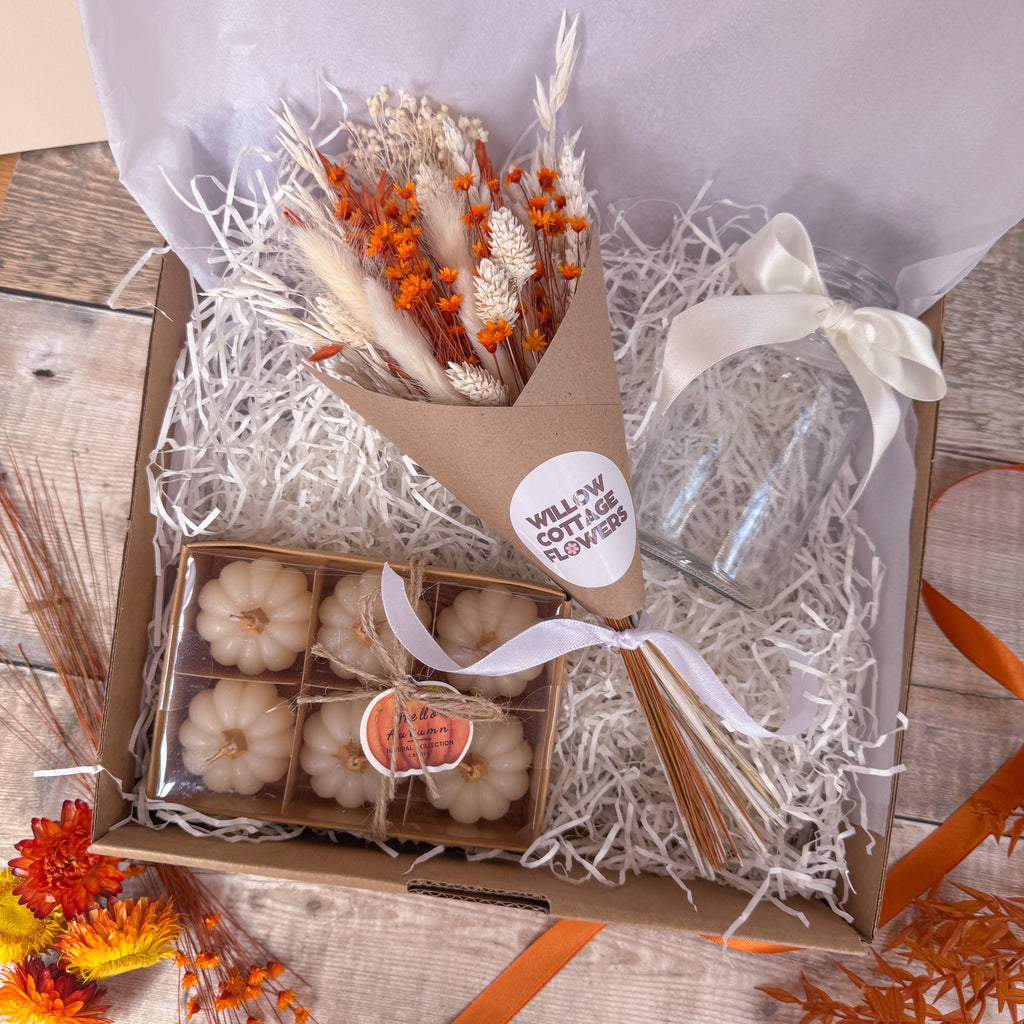 A gift box filled with white shredded paper contains a small bouquet of dried white and orange flowers wrapped in kraft paper, a clear glass vase tied with a white ribbon and a boxed set of small white pumpkin candles.