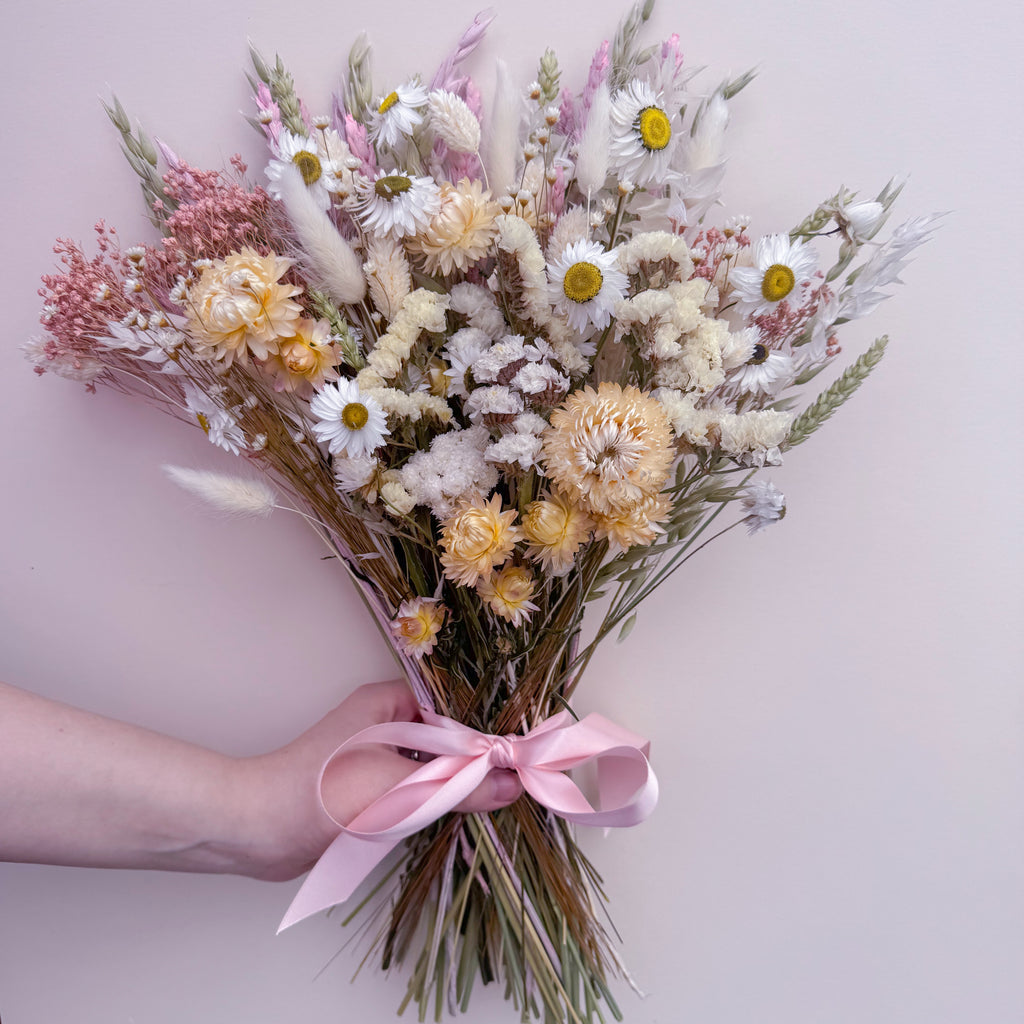 A hand holding a dried flower bouquet with white daisies, cream helichrysums, pink filler flowers, and bunny tails tied with a light pink ribbon, against a pale pink background.