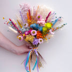 A hand holding a vibrant, multicolored dried flower bouquet with pastel and bright blooms tied with a blue, pink, and orange ribbon, set against a plain light gray background.