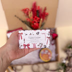 Hand holding a soap bar with decorative packaging featuring bows and text, with a blurred background of flowers and gift boxes.
