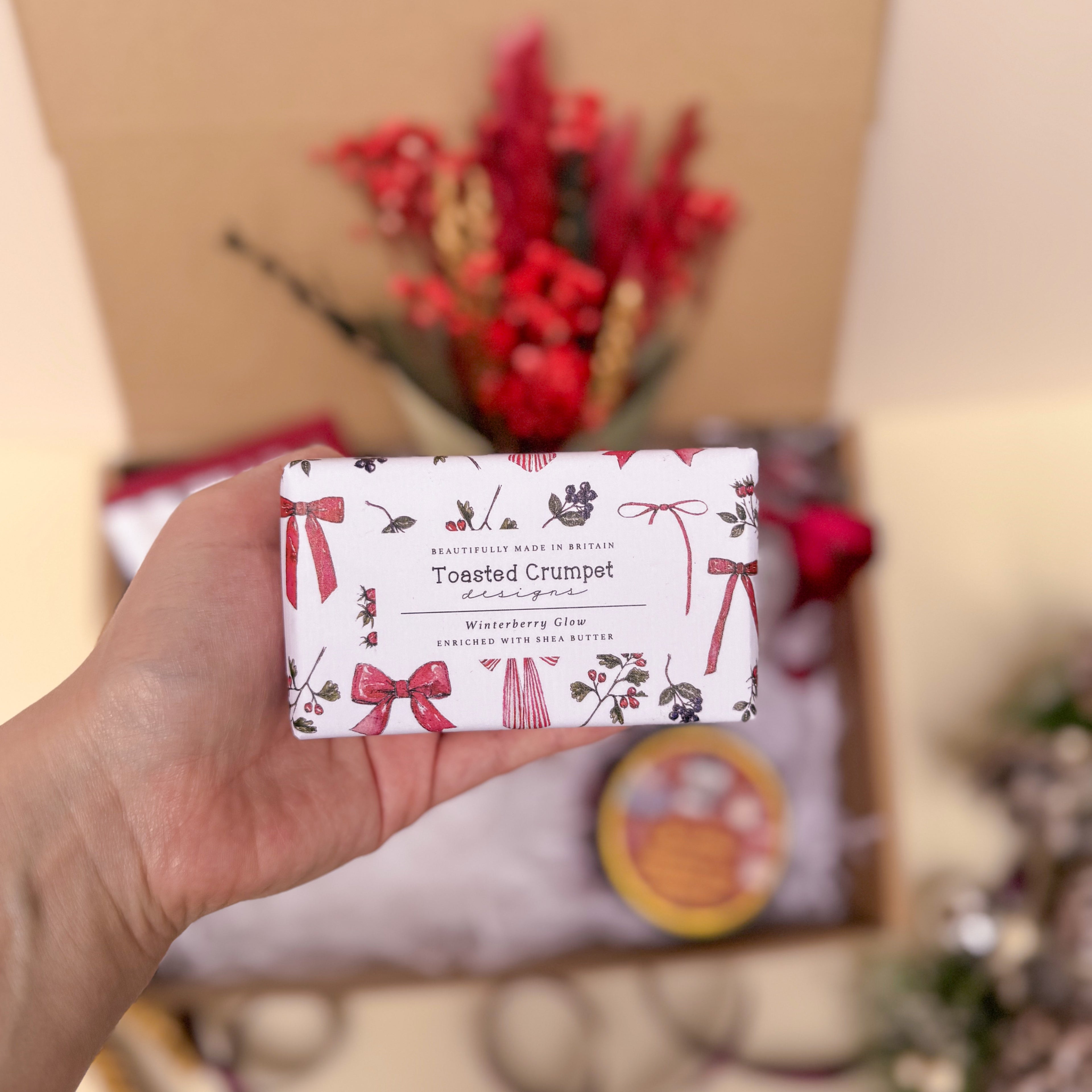 Hand holding a soap bar with decorative packaging featuring bows and text, with a blurred background of flowers and gift boxes.